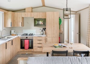 A modern kitchen with wooden cabinets, a built-in oven, and a dining area with a table and chairs. - Luxury 3 bedroom Hot Tub sleeps 8 Pet Friendly (Hayle)