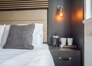 A neatly made bed with white and gray bedding, a gray ribbed pillow, a black nightstand with a coffee maker, two white cups, and a book titled 'Winchelsea' by Alex Preston. - Glamping Pods with hot tubs (Hayle)