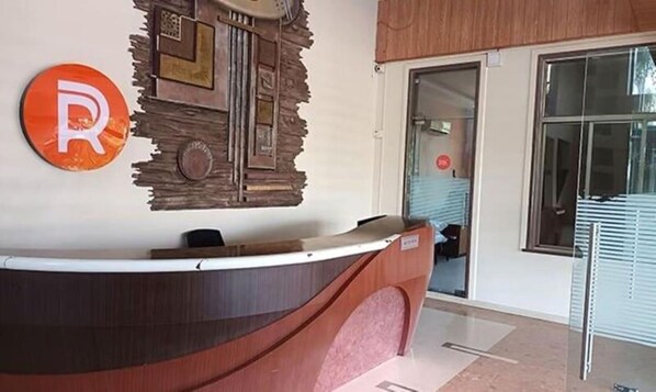 A reception desk with a wooden design, a large 'R' sign, and a glass door leading to a room. - Rester Select Chakan (Rajgurunagar)