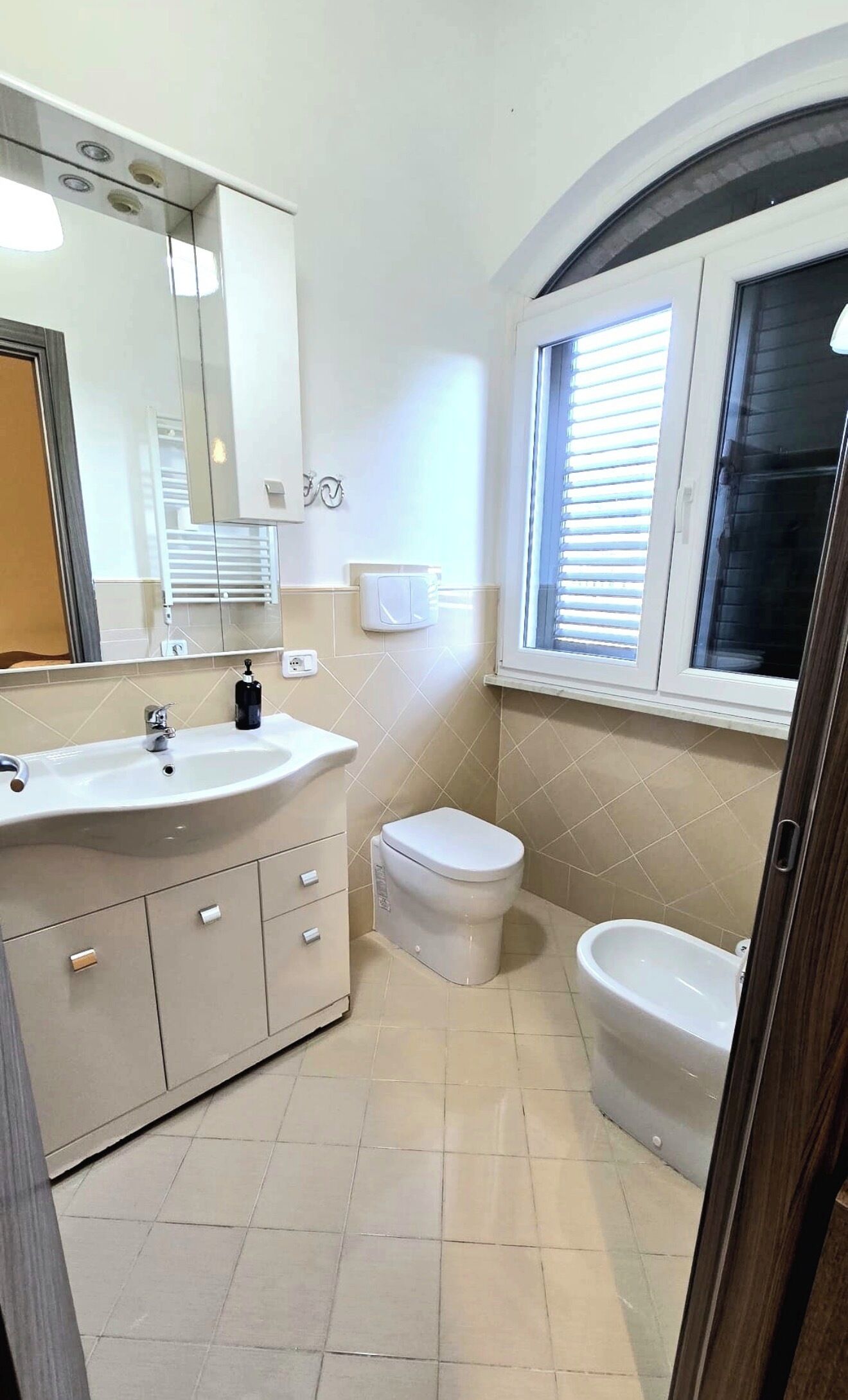 Ein Badezimmer mit Waschbecken, Toilette, Bidet und einem Fenster mit Jalousien.