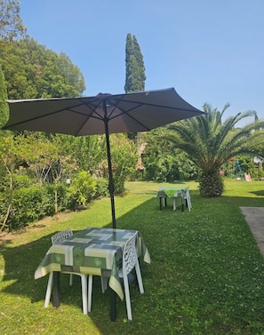 An outdoor seating area with a table and chairs under a large umbrella, surrounded by lush greenery and a tall tree.