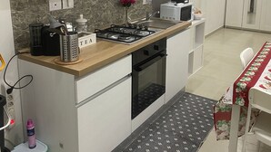A compact kitchen with white cabinets, a built-in stove, and a microwave. There's a small stool with a bottle on it, and a rug on the floor. - Giampy's house (Catania)
