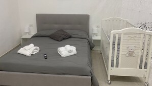 Een slaapkamer met een grijs bed, twee witte nachtkastjes met lampen, een wit ledikant met een teddybeer dessin en een aan de muur hangend schilderij.