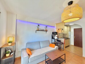 Ein Wohnzimmer mit einer Couch, einem Couchtisch, einem Fernseher auf einem Schrank sowie einer Küchenzeile mit Kühlschrank, Mikrowelle und Spüle.