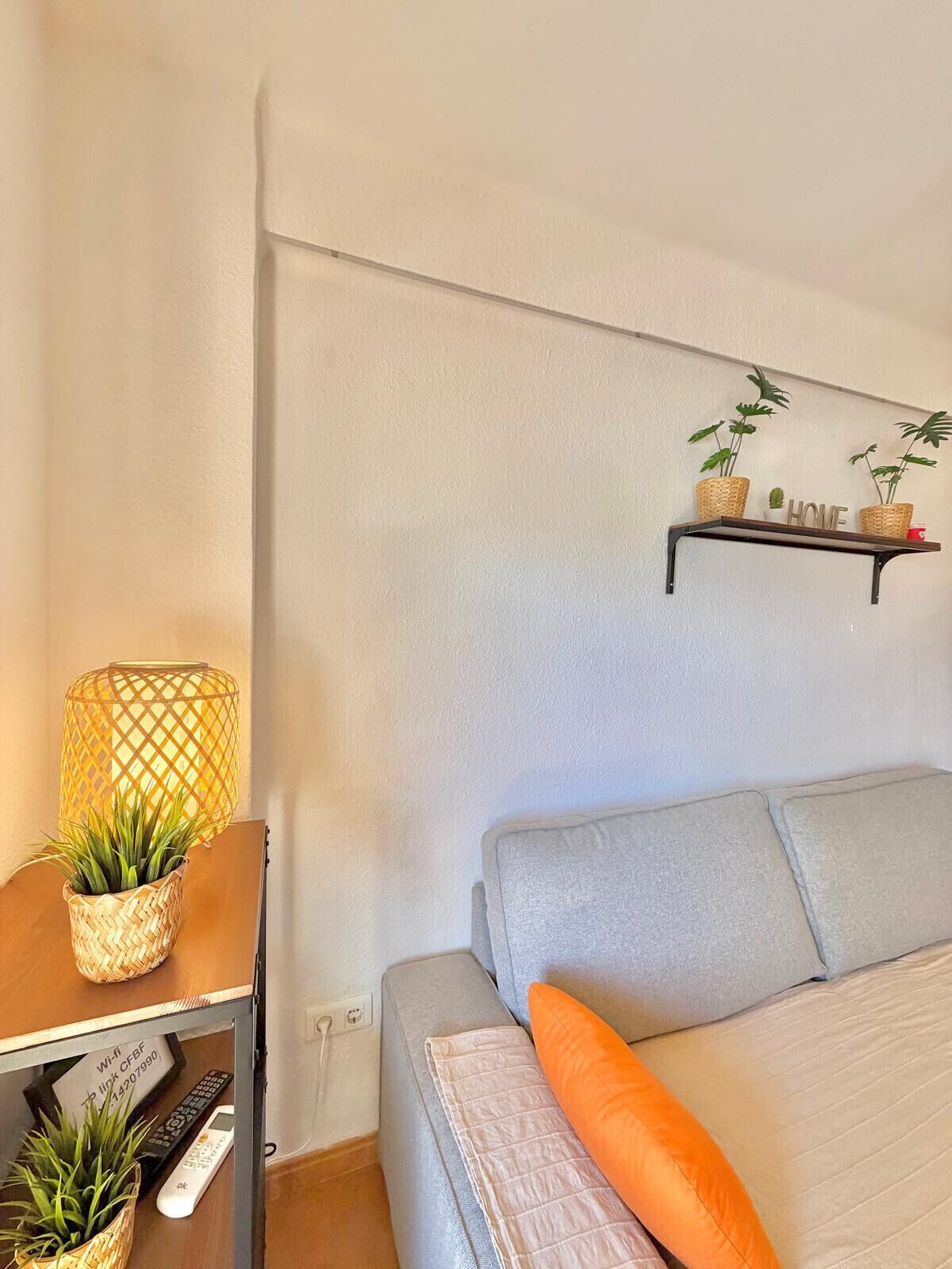 Een slaapkamer met een bed, een nachtkastje met een lamp, een plank met planten, en een bord waarop staat 'HOME'.