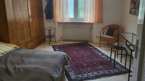 A bedroom with a large wardrobe, a bed, a chair, a small table, and a window with curtains. - Cozy garden apartment near Herrsching (Herrsching am Ammersee)