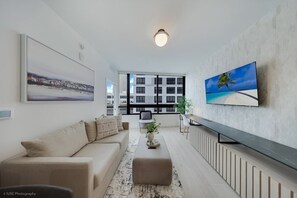 A modern living room with a sofa, a coffee table, a TV mounted on the wall, and a large window offering a view of buildings. - 225 DISTRICT LUXURY APARTMENT DOWNTOWN (MIAMI)