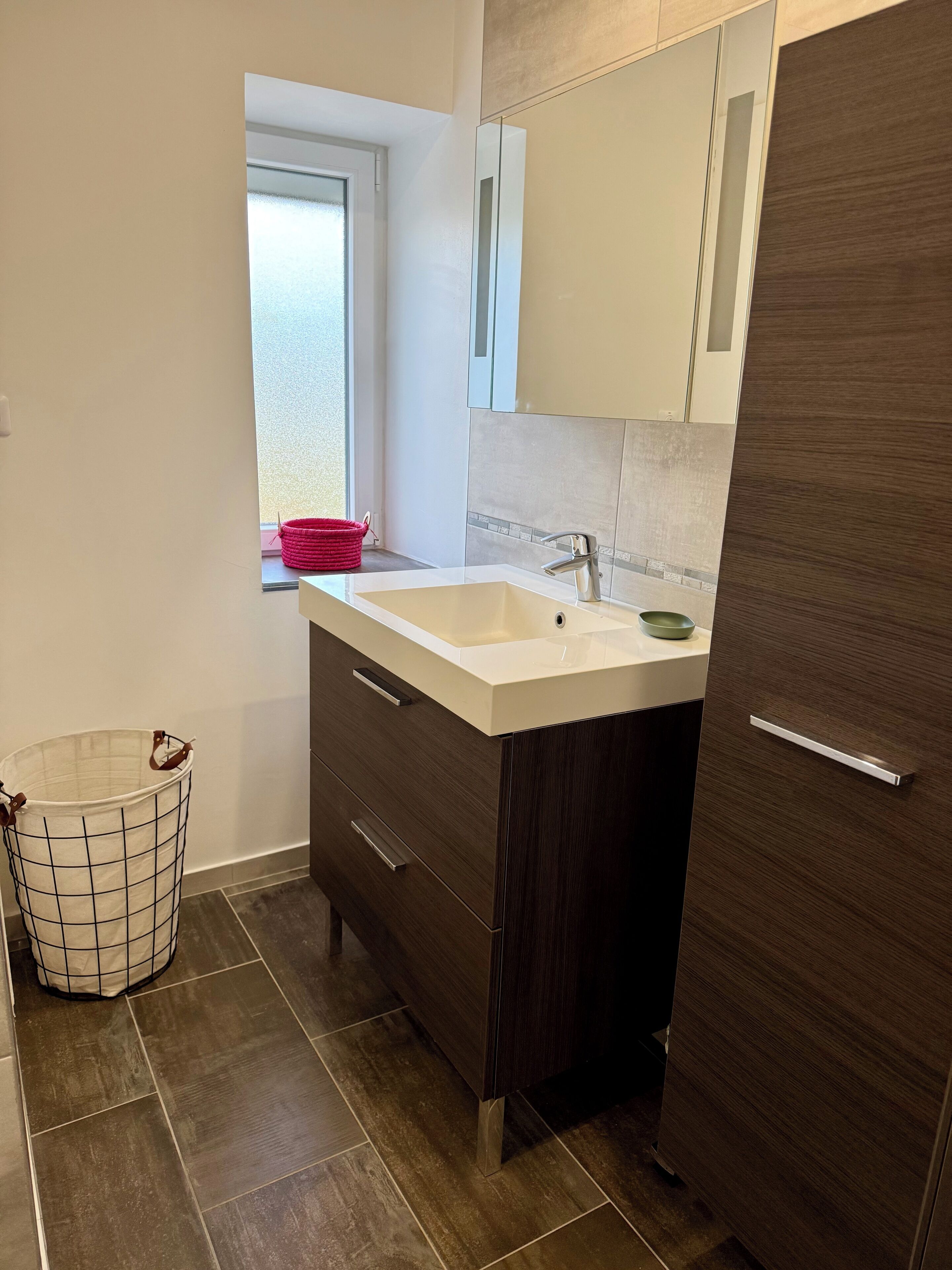 Ein modernes Badezimmer mit einem großen Waschbecken, dunklen Holzschränken, einem Spiegel und einem Fenster mit Blick.