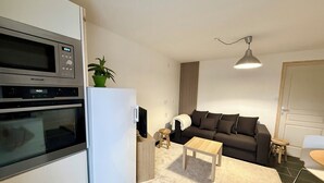 A compact kitchen with a Brandt oven, a white refrigerator, a small wooden table, a beige rug, and a dark brown sofa. - L'Aube - Cosy gîte with nature view for 4 people (Corcieux)