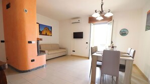 Ein Wohnzimmer mit beiger Couch, einem Esstisch mit Stühlen, einem an der Wand montierten Fernseher und einem Gemälde über der Couch.