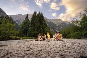 A group of people sitting around a campfire on a rocky riverbank with mountains in the background.