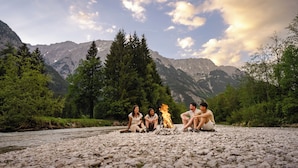 A group of people sitting around a campfire on a rocky riverbank with mountains in the background. - Relax in the Premium Room of Leutasch Retreat: extra space, bath and shower, ideal for active summers and winters in Tyrol. (Leutasch)