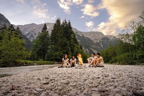 Un gruppo di persone sedute attorno a un fuoco da campo su una sponda rocciosa di un fiume, con montagne sullo sfondo.
