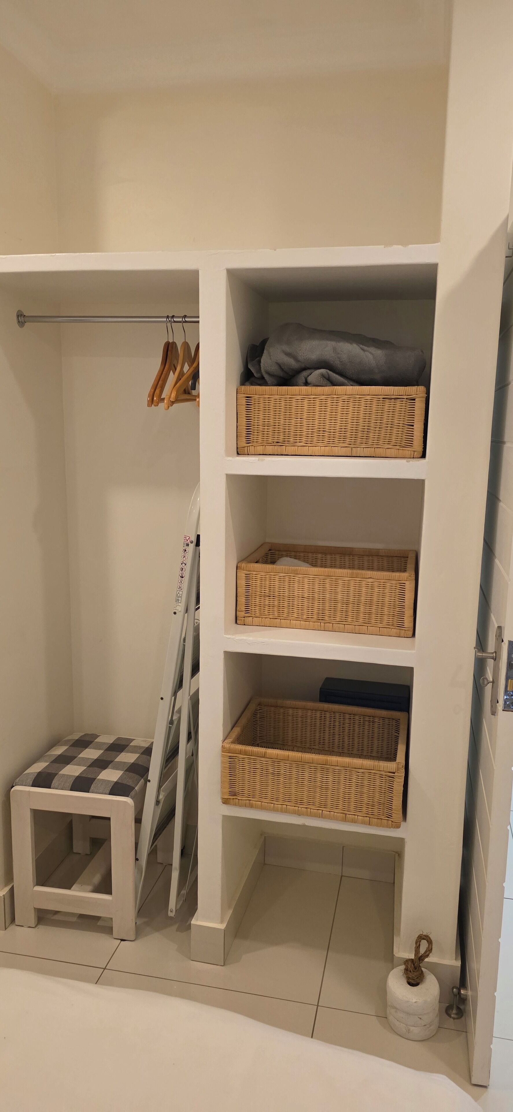 Un armario blanco con cestos de mimbre en estantes, un taburete pequeño y una barra metálica para colgar ropa.