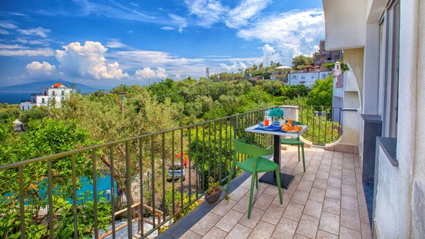 A balcony with a table and chairs, overlooking a lush green landscape and a distant view of buildings and a mountain. - Sorrento Sea View Villa - 4BR, Panoramic Terraces & Private Parking (Priora)