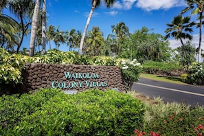 A sign for Waikoloa Colony Villas with a road leading to it, surrounded by lush greenery and palm trees. - 3BR Golf Course View Condo Waikoloa Colony Villas #805 - Vrkcv (Waikoloa)