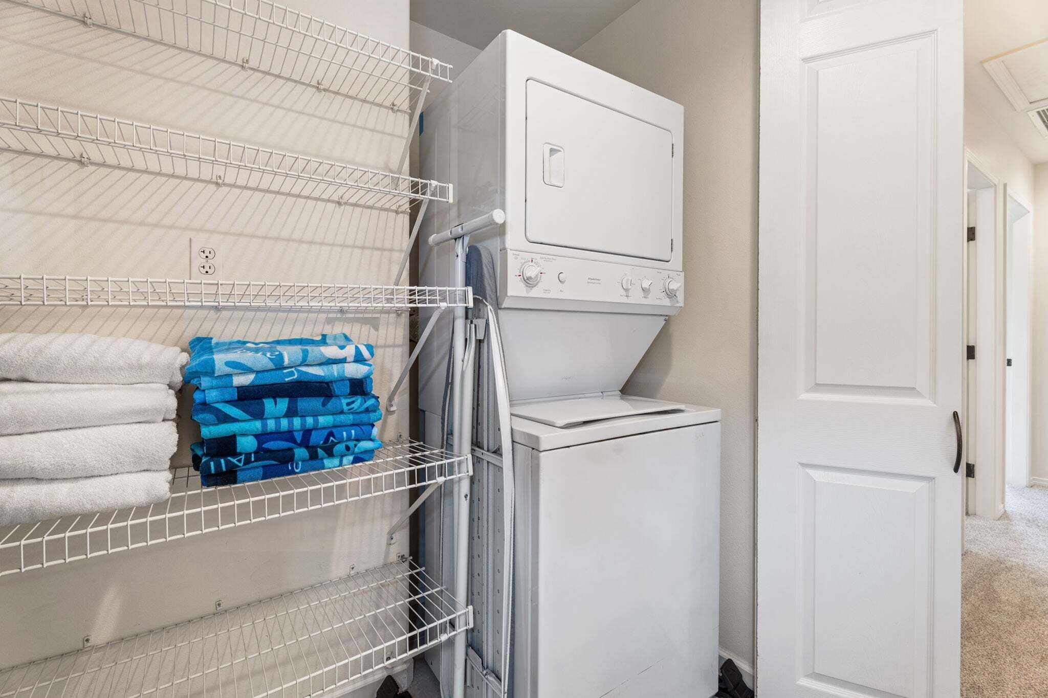 A laundry room with a stacked washer and dryer, metal shelving with folded towels, and a white door.