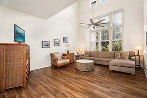 A spacious living room with a large window, a ceiling fan, and a flat-screen TV mounted on a wooden cabinet. - 3BR Golf Course View Condo Waikoloa Colony Villas #805 - Vrkcv (Waikoloa)