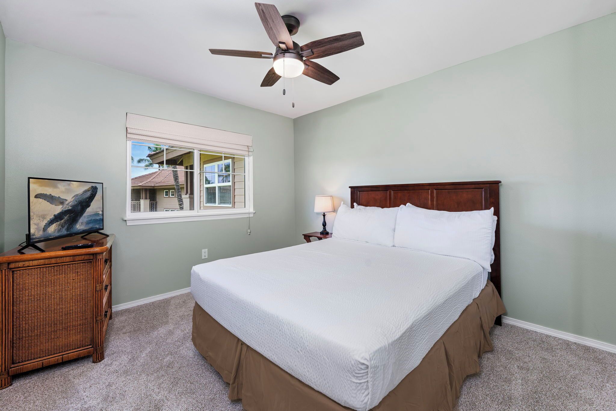 A bedroom with a bed, a ceiling fan, a window with blinds, a nightstand, and a wicker side table with a picture frame.