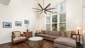 A living room with a sectional sofa, a round coffee table, a wicker armchair, a ceiling fan, and three framed pictures on the wall. - 3BR Golf Course View Condo Waikoloa Colony Villas #805 - Vrkcv (Waikoloa)