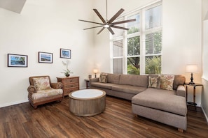 A living room with a sectional sofa, a round coffee table, a wicker armchair, a ceiling fan, and three framed pictures on the wall. - 3BR Golf Course View Condo Waikoloa Colony Villas #805 - Vrkcv (Waikoloa)