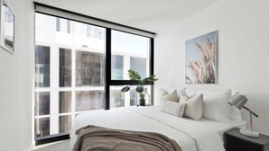 Ein modernes Schlafzimmer mit großem Fenster, einem Bett mit weißer Bettwäsche, einem Nachttisch mit Lampe und einem gerahmten Kunstwerk an der Wand.