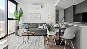 Ein modernes Wohnzimmer mit grauem Sofa, Couchtisch und Essbereich mit Stühlen. Es gibt ein großes Fenster, eine Pflanze und eine Küchenzeile mit schwarzer Arbeitsplatte.