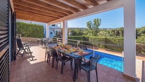 Un espace extérieur abrité, aménagé pour les repas, avec une piscine, des poutres en bois au plafond et une vue sur le paysage environnant.