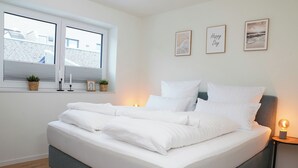 A neatly made bed with white linens, a grey headboard, and a bedside table with a lamp. There are three framed pictures on the wall and a window with blinds. - Küste33/8 - North Sea time on the dyke. Sauna. Beach chair. Balcony. (Tossens)