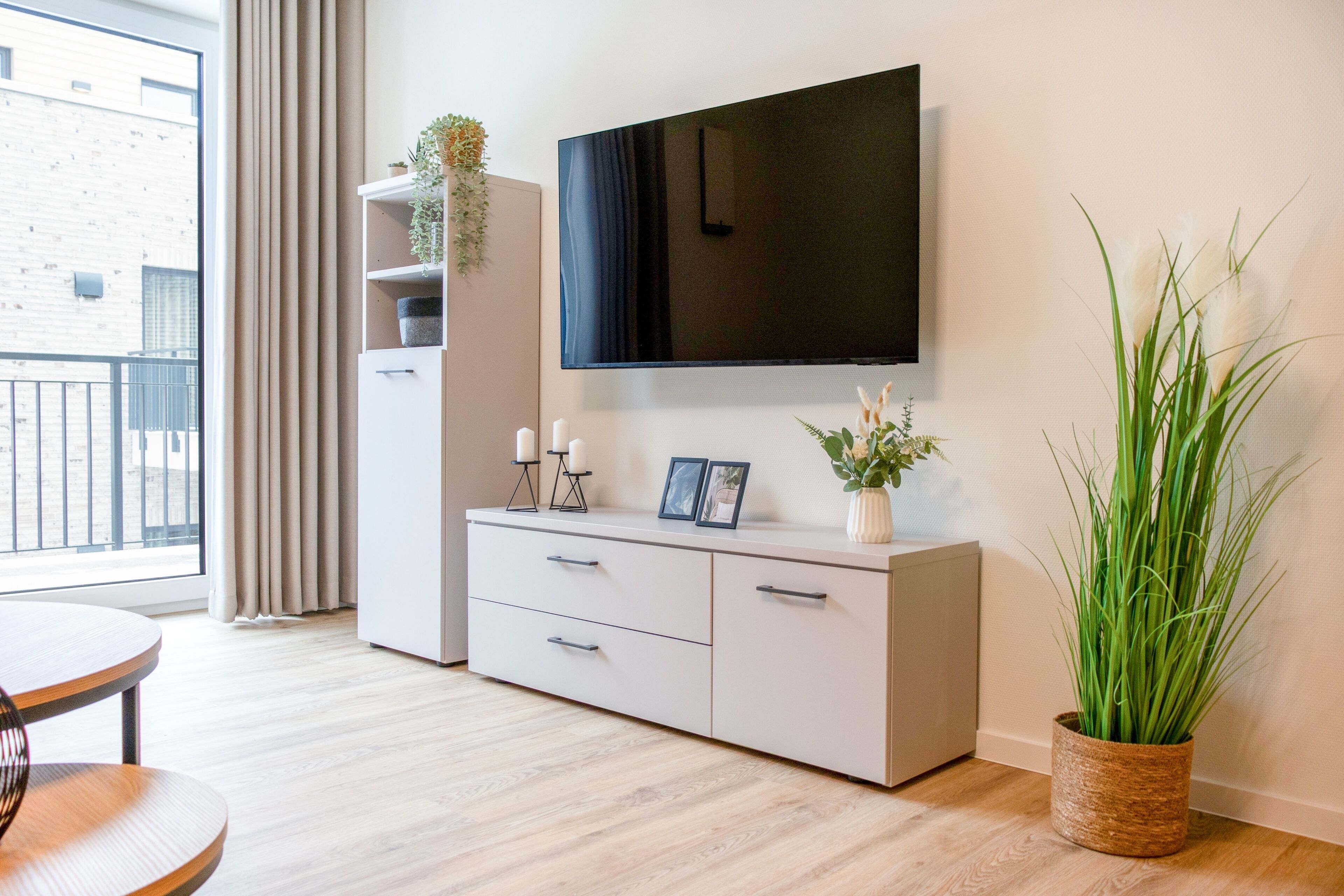 Un salon moderne avec un téléviseur à écran plat fixé au mur, un meuble blanc avec des tiroirs, une table ronde et une grande plante en pot.