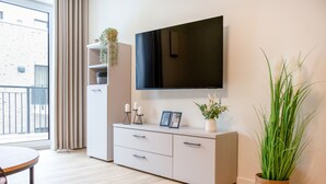 Un salon moderne avec un téléviseur à écran plat fixé au mur, un meuble blanc avec des tiroirs, une table ronde et une grande plante en pot.