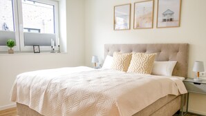 Un lit bien fait, recouvert d’une courtepointe, une table de chevet avec une lampe, une fenêtre avec des stores et des tableaux encadrés accrochés au mur.