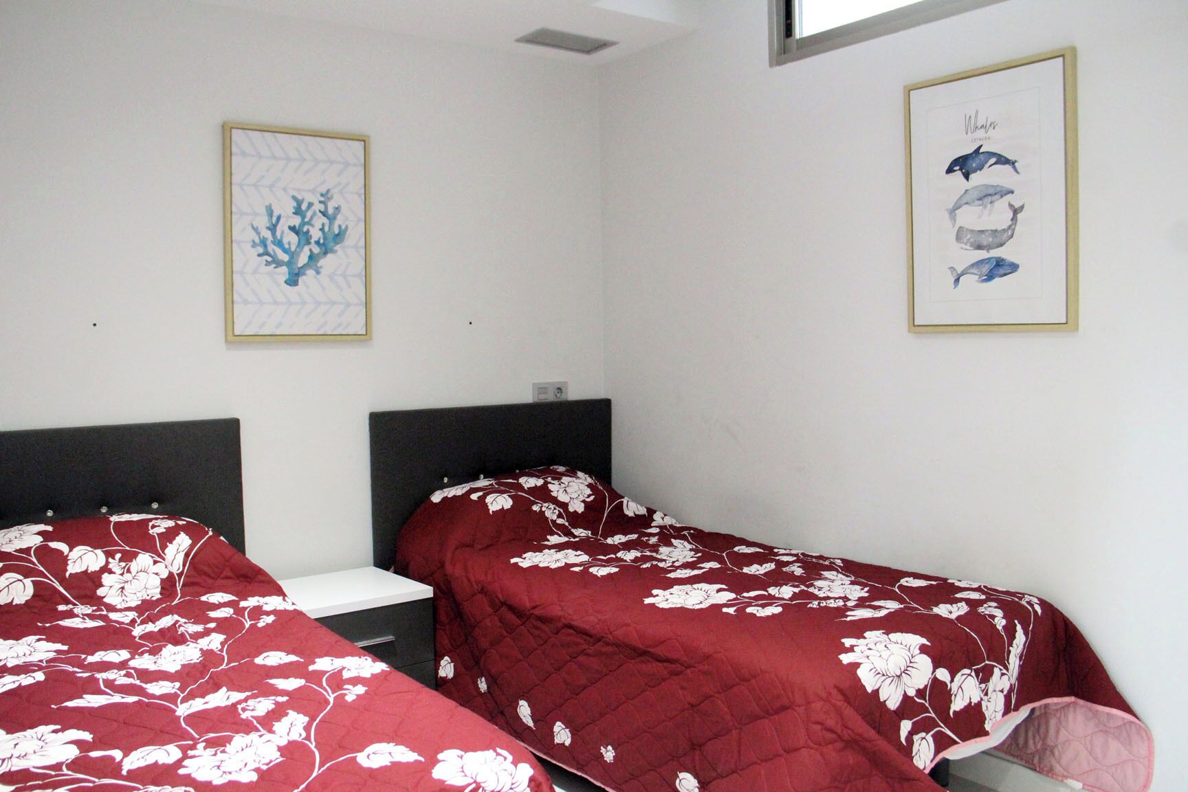 A room with two beds, each with a red floral bedspread, a white nightstand, and a framed picture of whales on the wall.