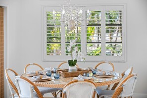 A dining area with a round table set for six, surrounded by wicker chairs, a chandelier, and a window with blinds. - Iris-istible Options by Beach Boutique Rentals (Anna Maria)