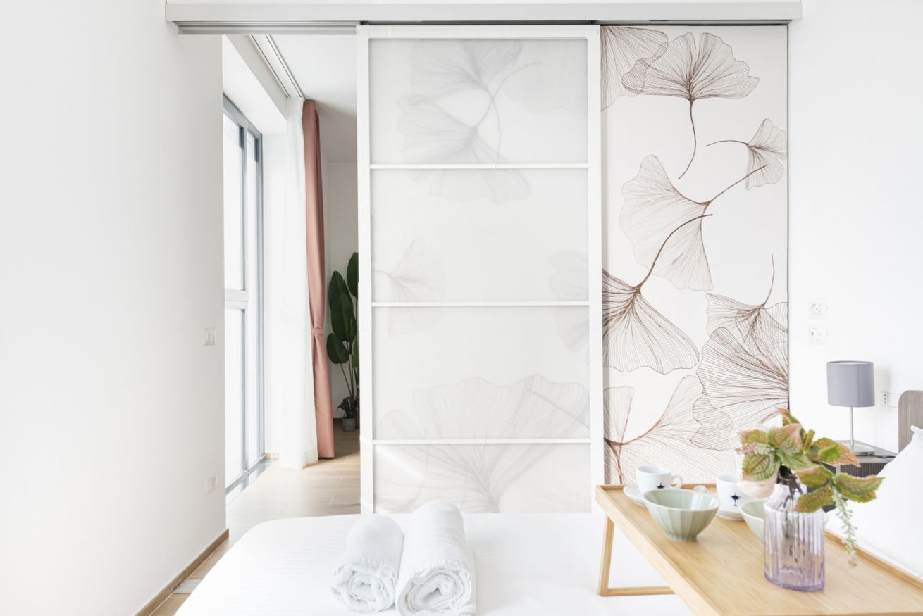 A modern hotel room with a large bed, a sliding door with a floral pattern, a bedside table with a lamp, and a view of a plant outside.