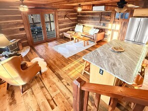A cabin interior with a wooden floor, a sofa, a dining table, and a kitchen area. - Bear Cove - rustic mountainside cabin with incredible view + free train tix (Bryson City)