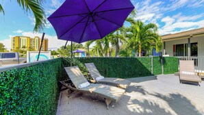 A poolside area with two lounge chairs, a purple umbrella, and a hedge wall. - A boutique, renovated coastal retreat featuring fully equipped condos w/kitchen, minutes walking to the beach & restaurants, exciting amenities like a heated pool, BBQ Grill, hammock and fun yard games. Centrally located, minutes to Pompano Beach! (Pompano Beach)