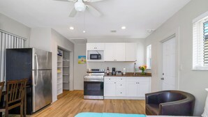 A compact kitchen with white cabinets, a stainless steel refrigerator, an oven, and a microwave. There is a ceiling fan, a window with blinds, and a wooden chair. - A boutique, renovated coastal retreat featuring fully equipped condos w/kitchen, minutes walking to the beach & restaurants, exciting amenities like a heated pool, BBQ Grill, hammock and fun yard games. Centrally located, minutes to Pompano Beach! (Pompano Beach)