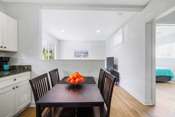 A modern kitchen with white cabinets, a dining table with a fruit bowl, and a TV in the background. - A boutique, renovated coastal retreat featuring fully equipped condos w/kitchen, minutes walking to the beach & restaurants, exciting amenities like a heated pool, BBQ Grill, hammock and fun yard games. Centrally located, minutes to Pompano Beach! (Pompano Beach)