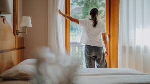 A person standing at a window with sheer curtains, a bed with white bedding, and a wooden headboard. - Waidmannhof (Merano)