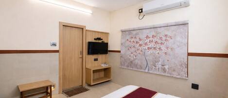 A hotel room with a bed, a TV mounted on a wooden cabinet, an air conditioner, and a wall-mounted artwork.
