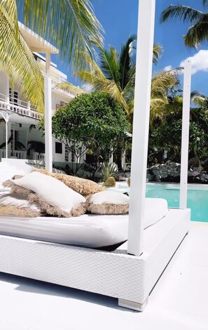 A white outdoor lounge area with a pool, surrounded by palm trees and a white building.