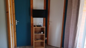 A room with a wooden door, a shelf with items, a television, and a trash bin.