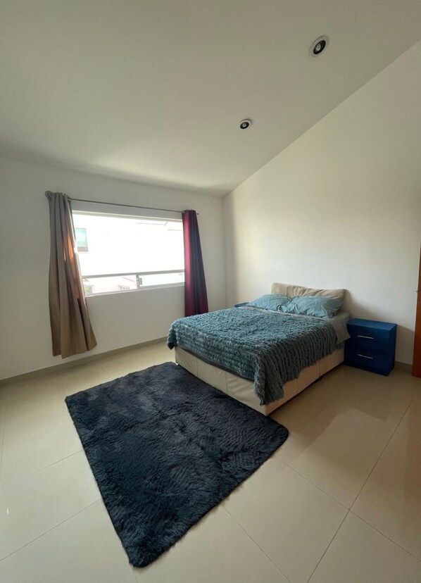 Une chambre à coucher avec un lit, un tapis foncé, une fenêtre avec des rideaux et une table de chevet bleue.