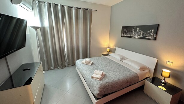A modern hotel room with a bed, a flat-screen TV, a nightstand with a lamp, and a cityscape painting on the wall. - 2 Bedroom Apartment Marconi Trastevere Station (Roma)