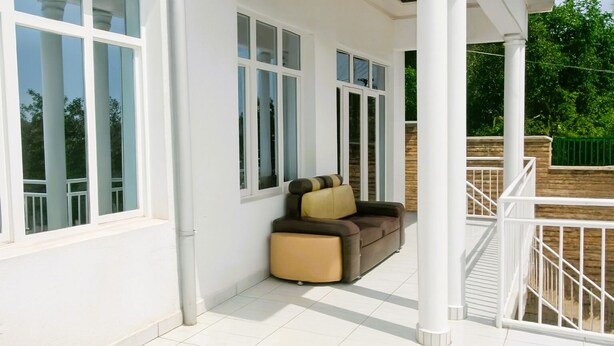 A covered outdoor seating area with a sofa and a chair, large windows, and a white railing. - Parkviewperch (Kigali)