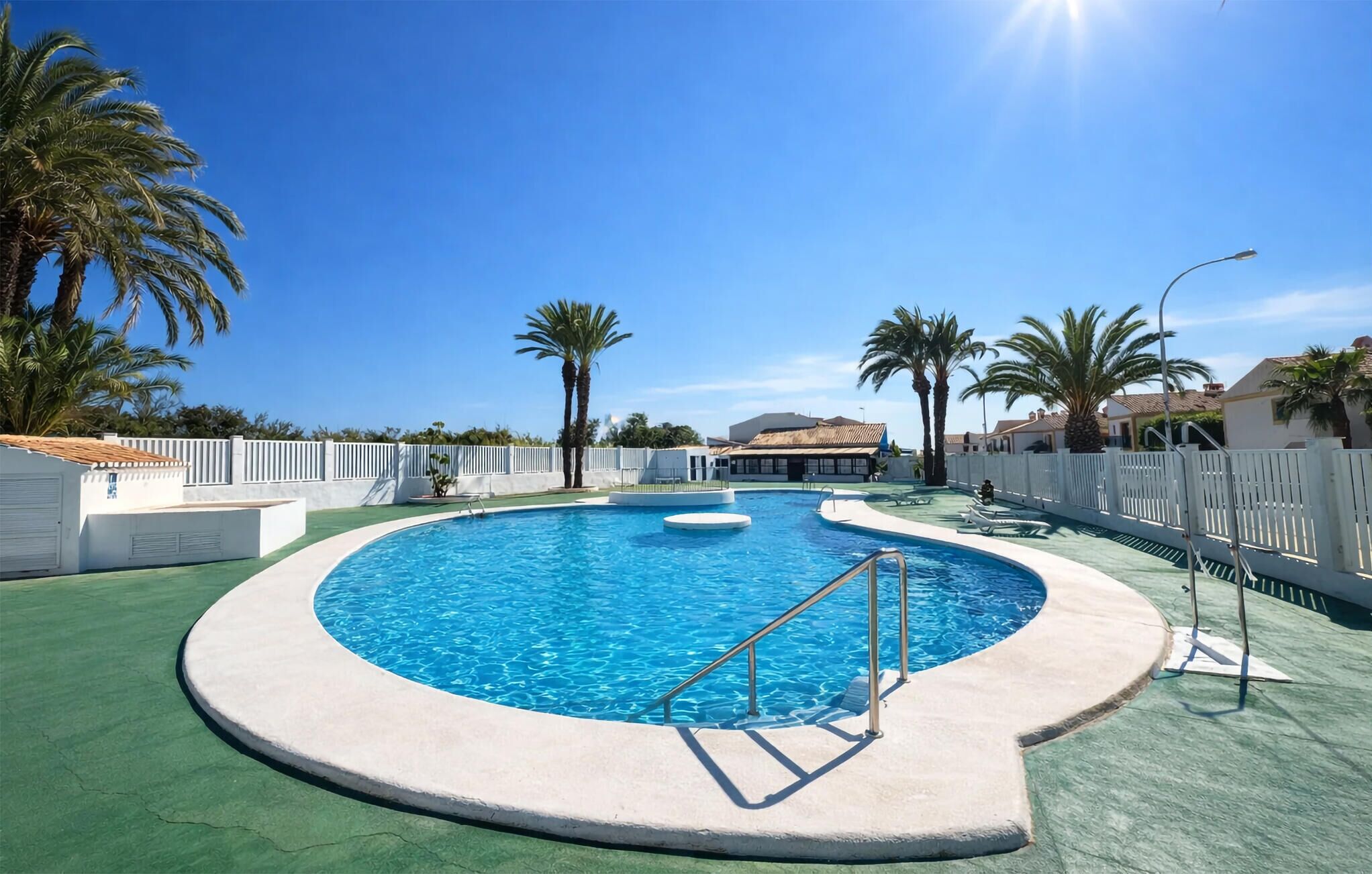 A circular pool with a diving board, surrounded by palm trees and a white fence.