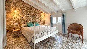 A bedroom with a stone wall, a bed with white metal frame, patterned floor tiles, a chair, and a window with curtains. - Lovely home in Petra with private swimming pool, can be inside or outside (Petra)