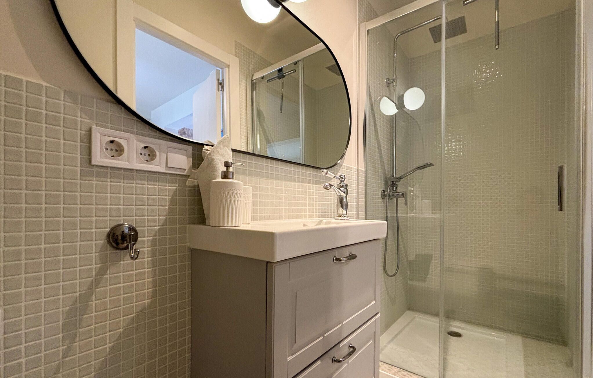 A bathroom with a tiled wall, a sink with a mirror, a towel dispenser, and a shower area.