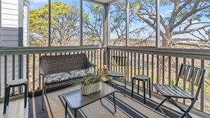 Un espace extérieur aménagé, abrité et doté d’un canapé, de chaises et d’une petite table, offrant une vue sur un paysage arboré et un bâtiment.